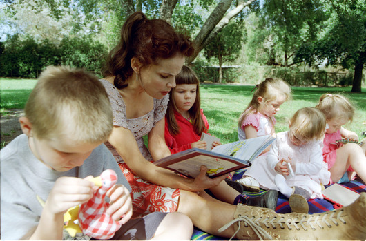 Sigrid and five children sit on picnic blanket as she reads a story, with two open suitcases beside them