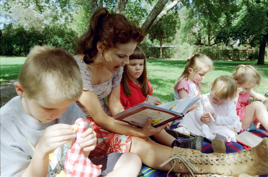 Sigrid and five children sit on picnic blanket as she reads a story, with two open suitcases beside them