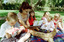 Sigrid and five children sit on picnic blanket as she reads a story, with two open suitcases beside them
