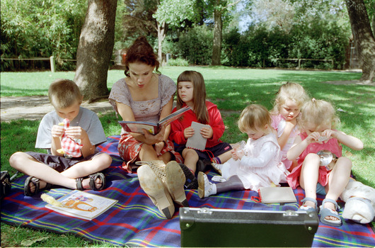 Sigrid and five children sit on picnic blanket as she reads a story, with two open suitcases beside them