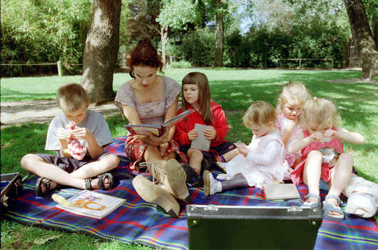 Sigrid and five children sit on picnic blanket as she reads a story, with two open suitcases beside them