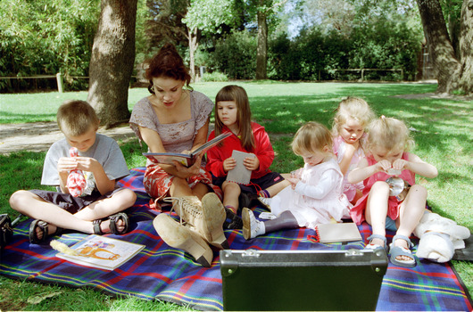 Sigrid and five children sit on picnic blanket as she reads a story, with two open suitcases beside them