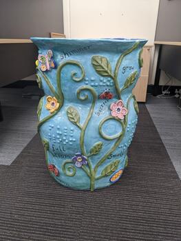 Light blue pot with vines, leaves, flowers, insects and words written in cursive and braille
