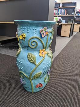 Light blue pot with vines, leaves, flowers, insects and words written in cursive and braille