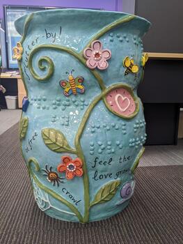Light blue pot with vines, leaves, flowers, insects and words written in cursive and braille