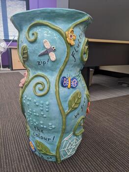 Light blue pot with vines, leaves, flowers, insects and words written in cursive and braille