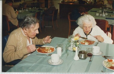 Two women sit at a table eating stewed meat and vegetables for lunch
