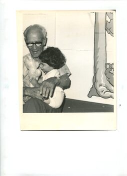 Older man holds young girl in his lap as she plays with a teddy bear