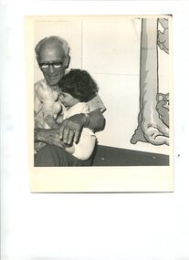 Older man holds young girl in his lap as she plays with a teddy bear