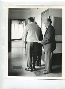 Three man stand in a corridor, one holding the jumper of the one before him and the other possibly an arm