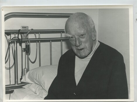 Older man sits on the side of a bed facing towards camera