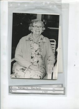 Older female dressed in cardigan and floral dress sits outside on an outdoor chair