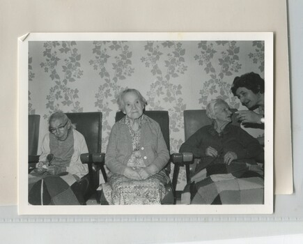 Three older women sit in lounge chairs along a wall with leaf wallpaper