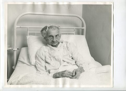 Elderly woman sits upright in hospital bed in her nightgown.