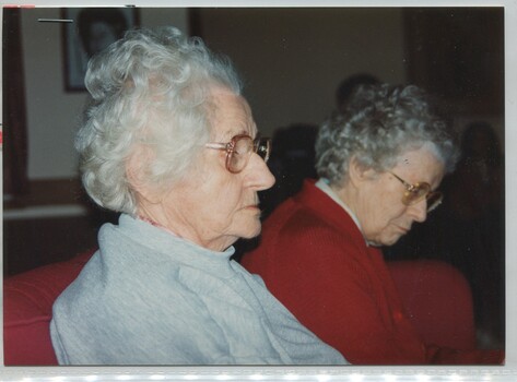 Two women sit on a red couch, one wearing a blue gray marle jumper and the other a red top