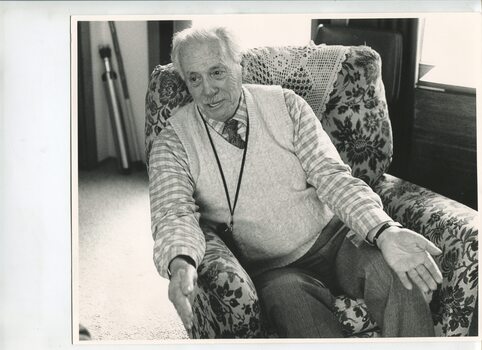 Man sit in a floral armchair with arms forward and hands apart mid story telling
