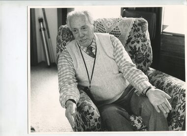 Man sit in a floral armchair with arms forward and hands apart mid story telling