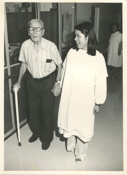 Elderly man holds his stick in one hand and a nurse's hand in the other, as he moves along the corridor