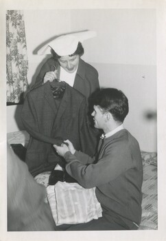 Matron Agar showing a new suit to Alex who sits on a bed with pyjamas and shoes in his lap