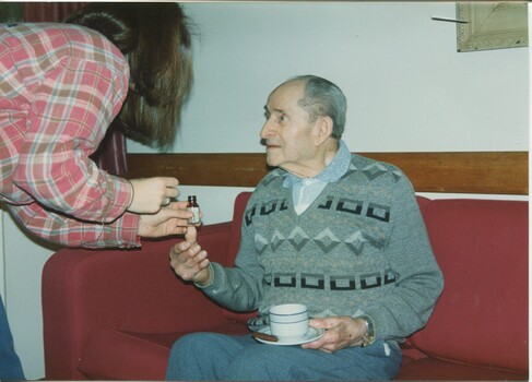 Older man balances a cup of tea on his lap as a woman leans towards him, holding a small bottle