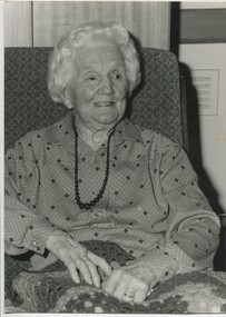 Older woman wearing string of beads and dress sits in a chair