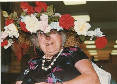 Woman smiles from under a hat of red, yellow, blue and pink fake carnations