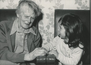 Young girl places her hand on to those of an elderly woman as they sit on lounge room chairs