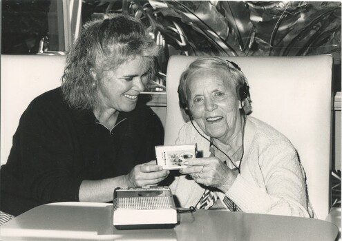 Older woman smiles as she listens to a tape, as a younger woman hands her the cassette case.