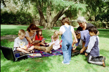Sigrid and various children sit on or stand beside a picnic blanket 