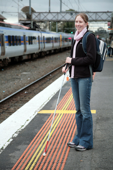 Anna waiting on the platform at a train station with her cane