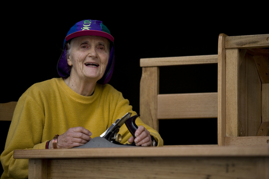 Elderly woman sits at a wooden table with planer in her hand