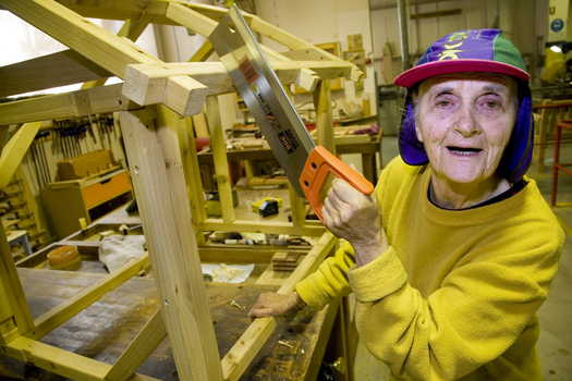 Elderly woman stands in a workshop with a saw in her hands