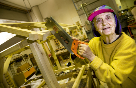 Elderly woman stands in a workshop with a saw in her hands
