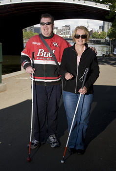Man and woman stand with their canes against St Kilda Road bridge and Yarra River backdrop