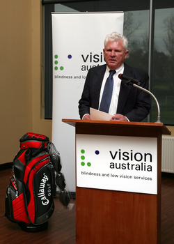 David Blyth speaking at the podium with his golf clubs beside him