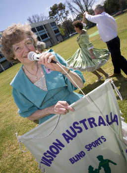 Margaret Fox with microphone and banner in front of square dancers at rear of Kooyong building
