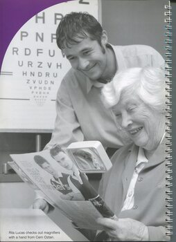 Rita Lucas using a magnifier to read a magazine whilst Cem Oztan stands beside her