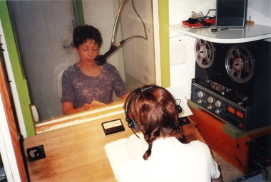 Lise Rodgers reads a book into a microphone whilst Carmel McAloon sits outside the booth, listening to the recording