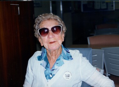 Elderly woman wearing white round badge with 'Mrs Edna Adams' in centre, 'Kooyong' at base and 'Association for the Blind' around the edge