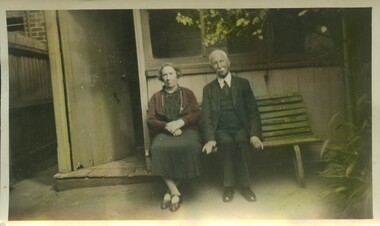 An elderly man and woman sit on a bench next to a shed in a backyard