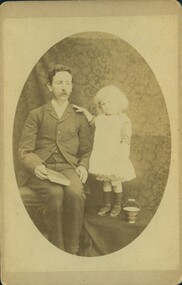 A man seated next to a girl, who has her hand on his shoulder, and she stands on a covered box