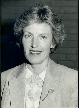 Woman wearing suede jacket, diamond patterned jumper and white shirt, and tennis racquet pin on lapel