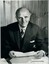 Man leans forward at desk, wearing a three piece suit, tie and kerchief in pocket