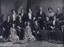 13 people pose for photograph in late 19th century/early 20th clothing