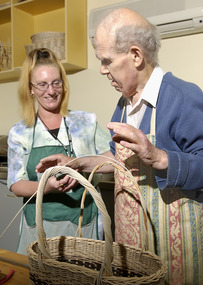 Woman holds a man's arm as he moves to bend a cane strip to make a basket handle.