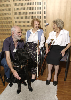 Two seated women in the Kooyong Day Centre with their walking sticks with Colin Pascoe and his dog