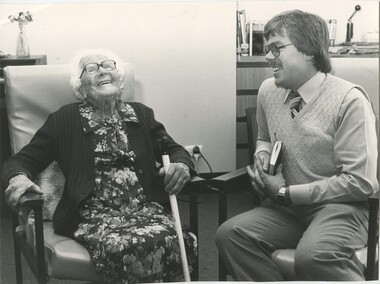 Elderly woman laughs with younger male as they sit in a lounge