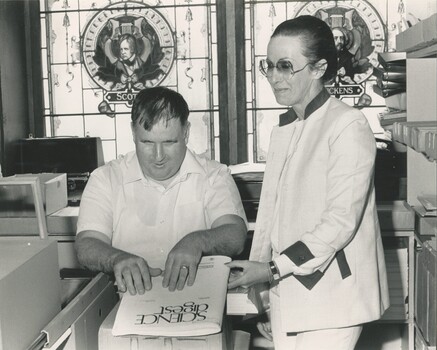 Man reading cover of Braille magazine, with lady beside him and stained glass windows in background