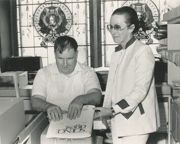 Man reading cover of Braille magazine, with lady beside him and stained glass windows in background