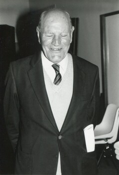 Older man in suit jacket, knit vest, white shirt and tie smiling towards camera